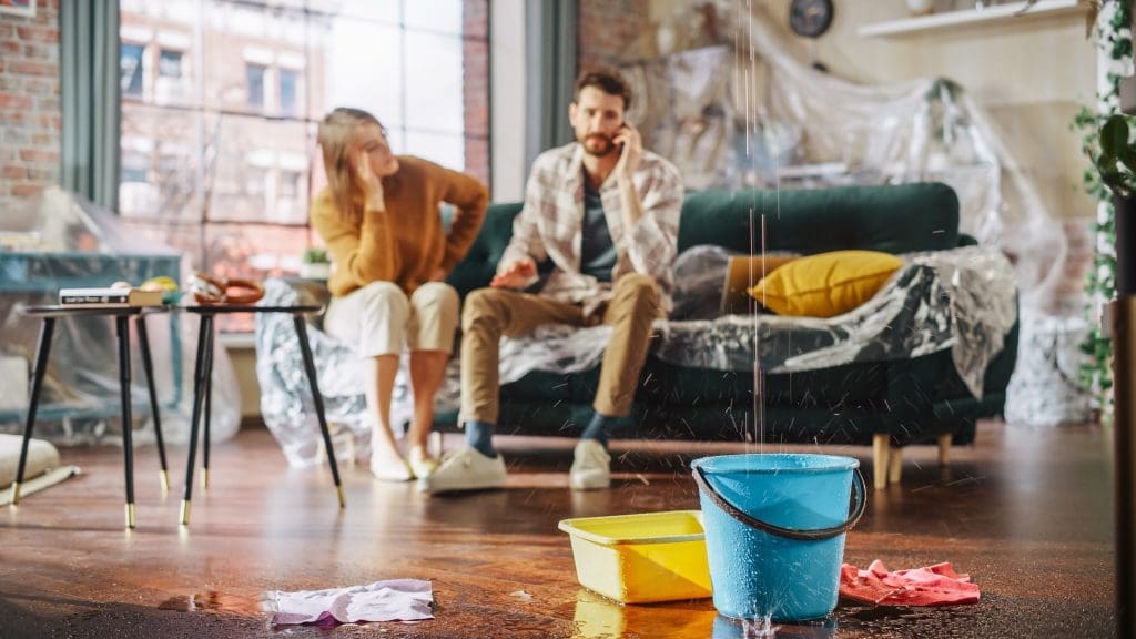 Couple sitting on couch in home while staring at water fill up in bucket as it leaks from the roof