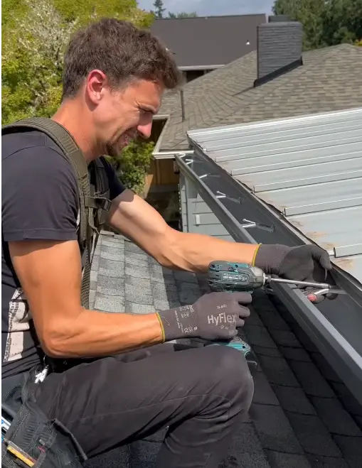 Professional from a roofing company in Monroe County using a drill to fasten a seamless gutter along a shingle roof.