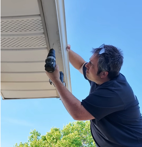 A roofing specialist from Daniel Hood Roofing installs new gutters on a home using a power drill under clear blue skies, showcasing expert gutter replacement in Knoxville, TN.